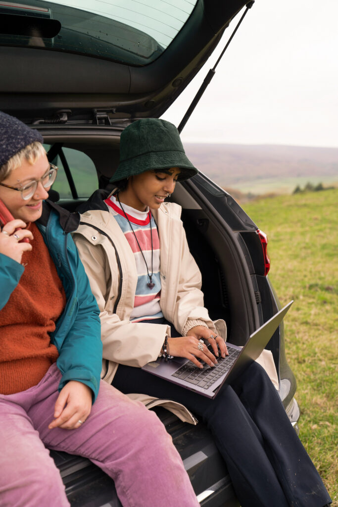 Female hikers sitting in car boot and using phone and laptop